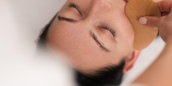 From above of crop unrecognizable beautician massaging face of calm young woman, lying on couch with closed eyes with wooden gua sha in spa salon