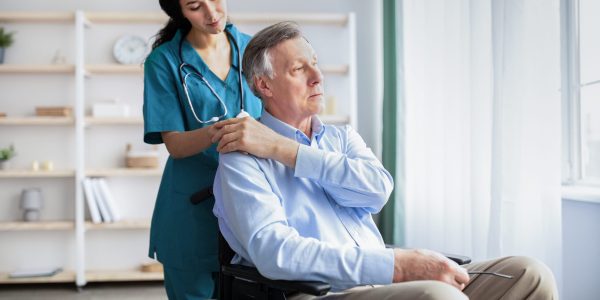 Young doctor holding elderly disabled male's hand, giving him support at retirement home. Senior man in wheelchair suffering from depression, millennial nurse helping him, indoors
