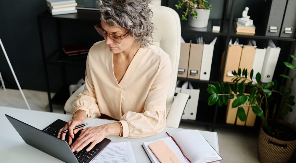 Mature female analyst or broker typing on laptop keyboard while sitting by workplace with financial documents, smartphone and open notepad