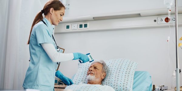 Hospitalized elderly man getting his temperature measured with infrared thermometer by nurse during his recovery.